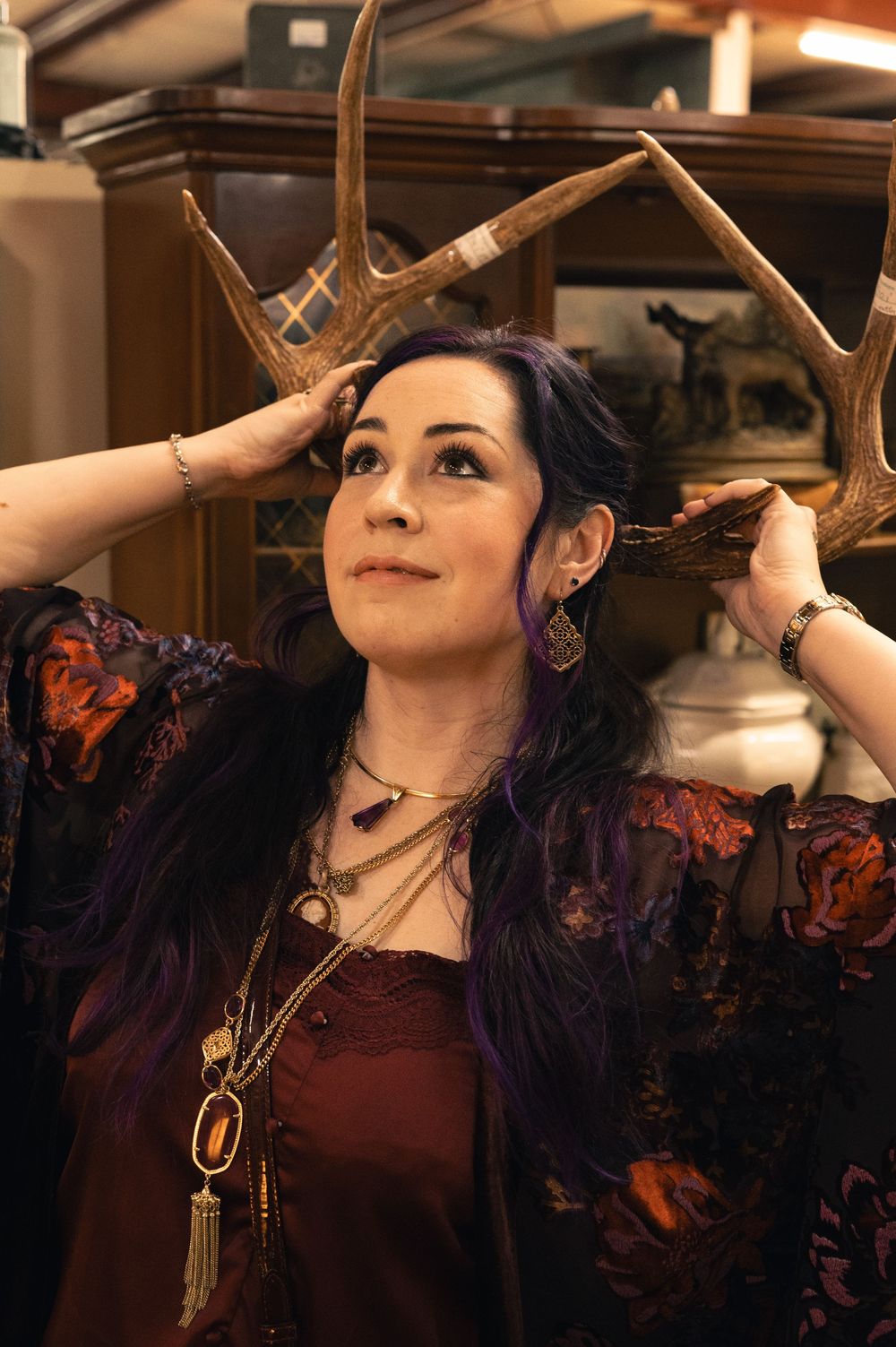 Woman with purple hair holds deer antlers above her head, wearing layered necklaces and a floral kimono.