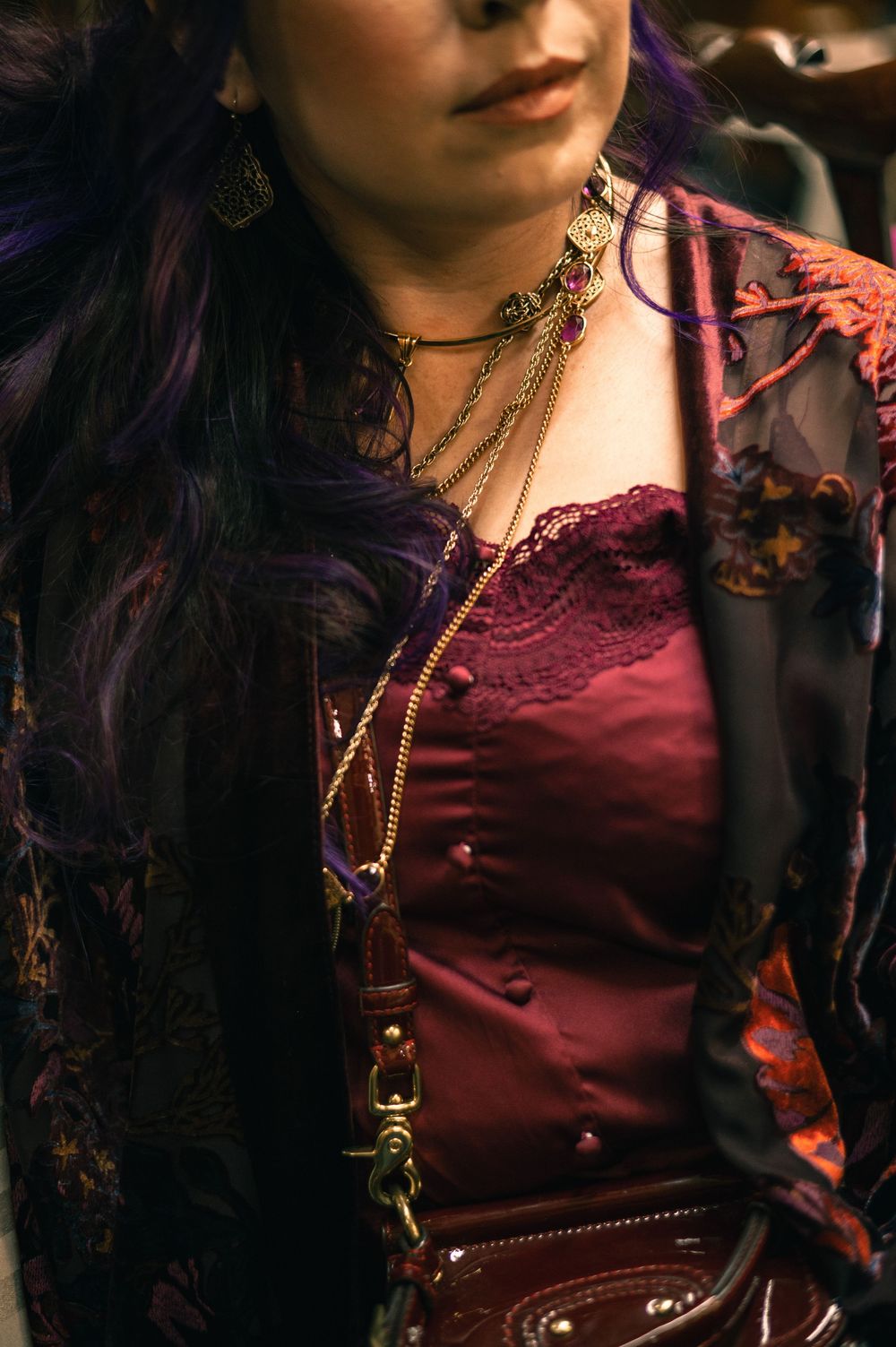 Woman in burgundy lace top with purple hair, layered gold chain choker necklace, and dark floral jacket.