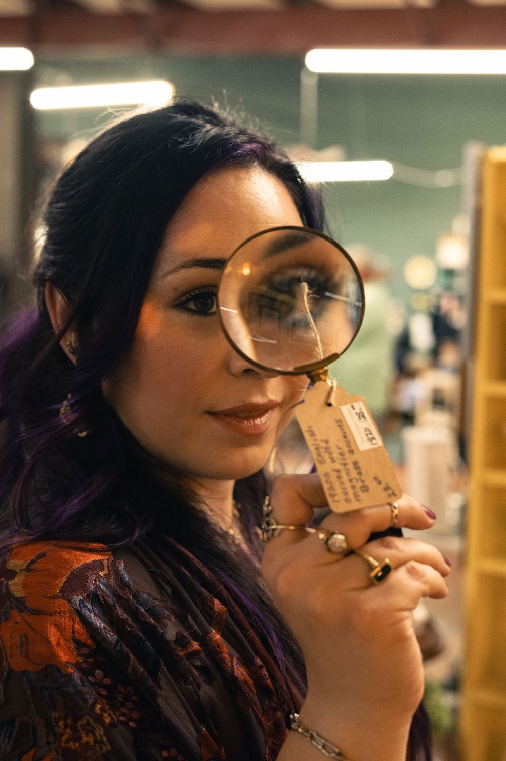 Woman with purple highlights holds magnifying glass to eye while examining antique item in shop.