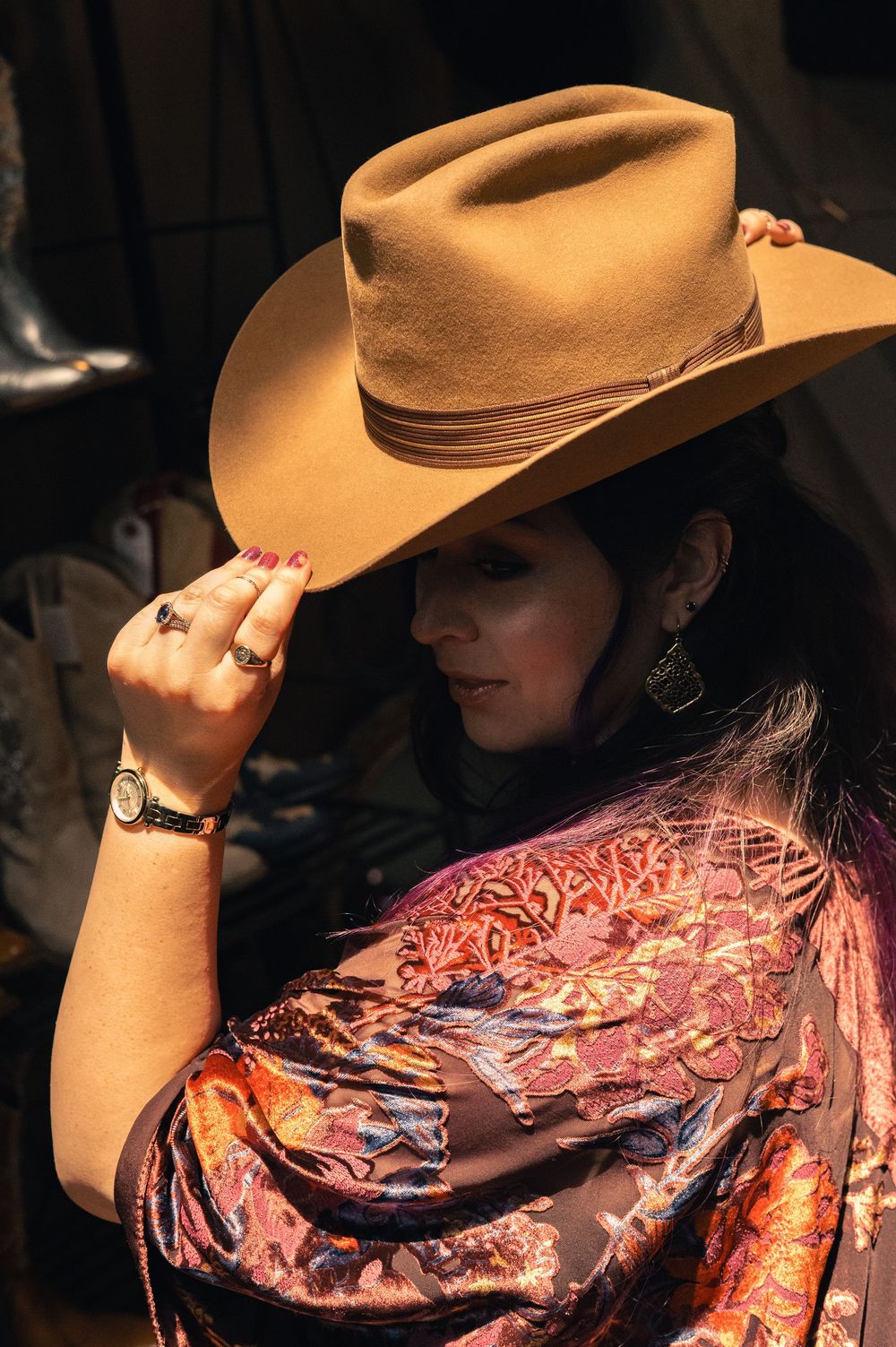 Woman in tan cowboy hat and floral shawl, adjusting brim with red-nailed hand, wearing gold watch and earrings.