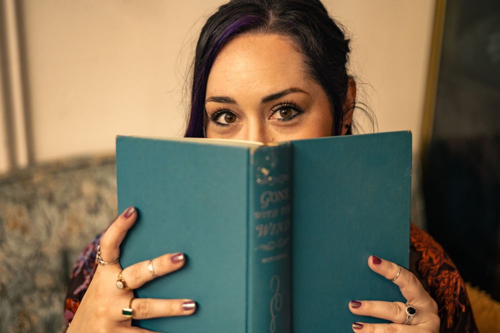 Woman with dark hair and rings holds a teal hardcover book up to her face, peering over the top with expressive eyes.