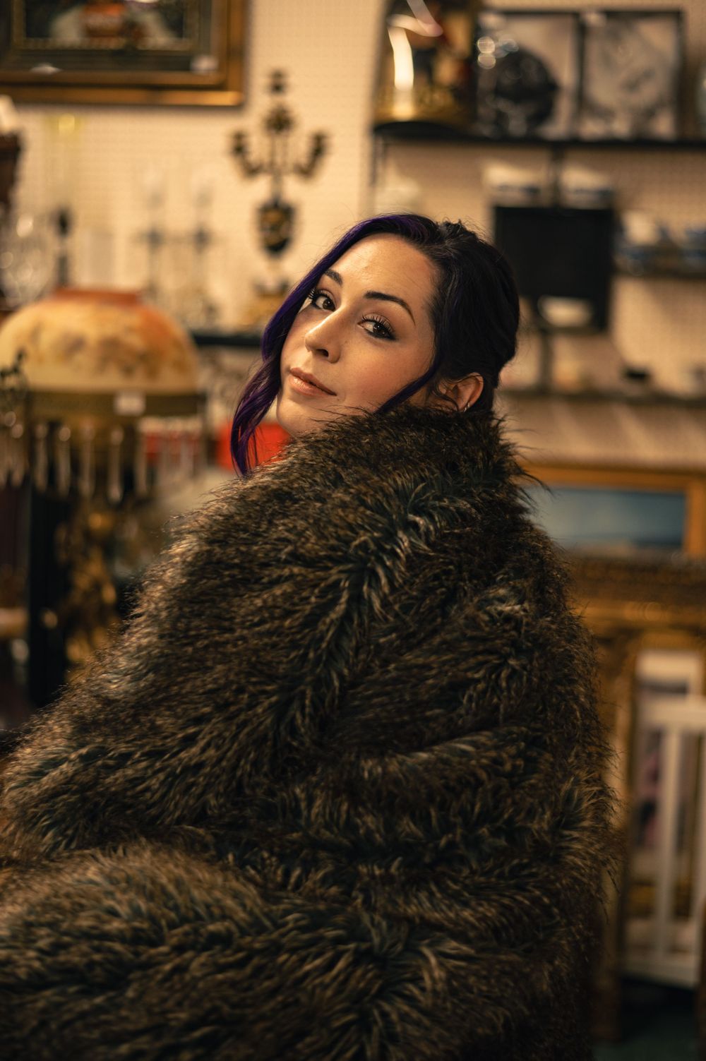 Young woman wrapped in a large fur coat posing in an antique shop with vintage decor and artwork.