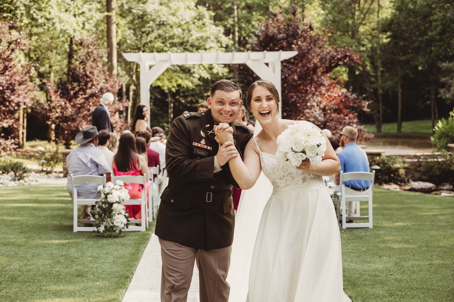 Happy newlywed couple celebrating outdoors after wedding ceremony, bride in white gown holding bouquet, groom in military dress uniform.