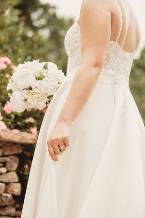 Bride in white lace wedding gown holding white peony bouquet with ring visible, standing near stone wall and pink roses.