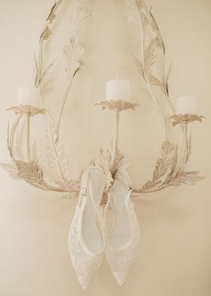 Elegant white lace bridal heels hanging on a vintage cream metal wall sconce with candles and leaf details.