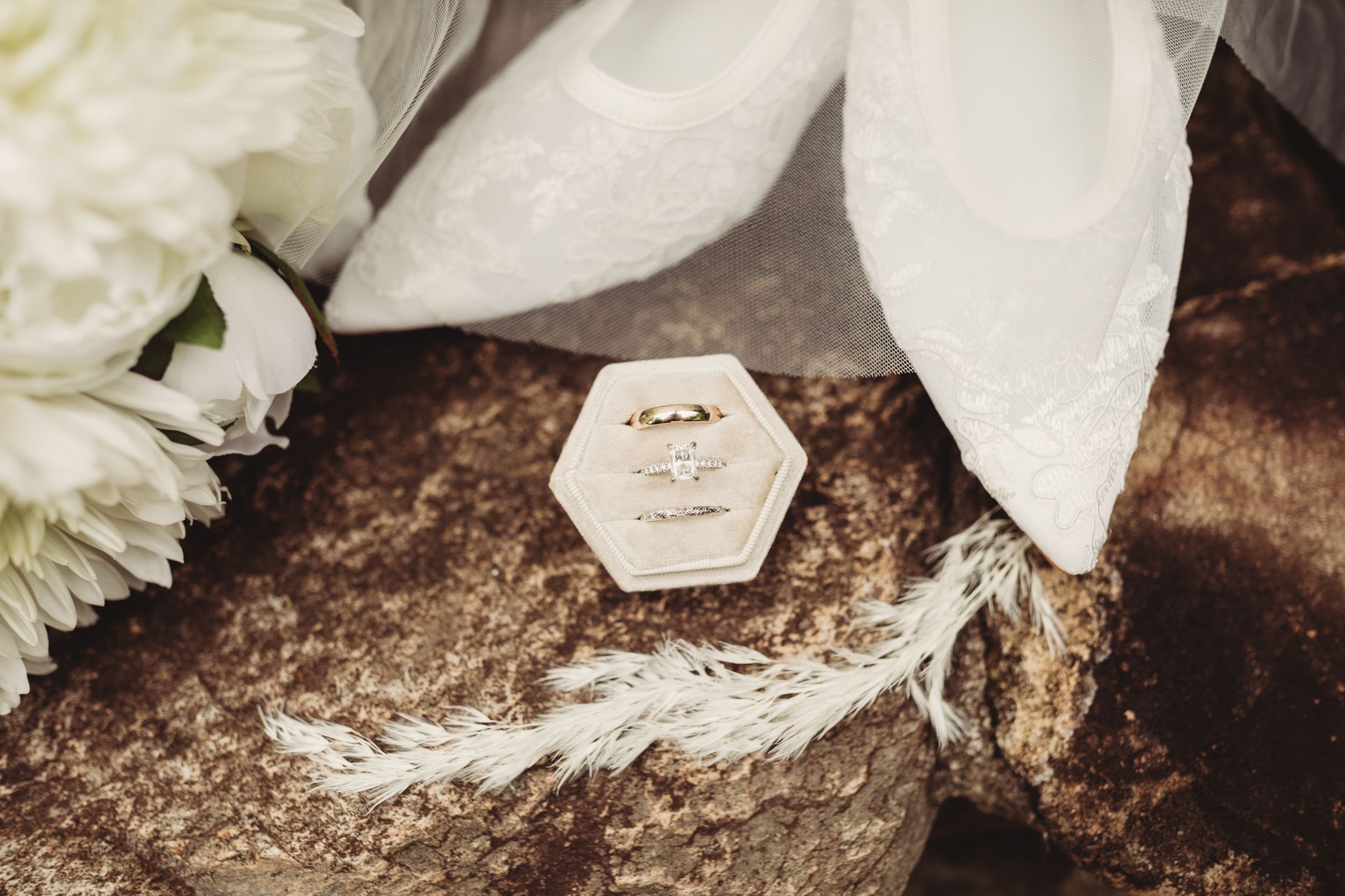 Elegant hexagonal velvet ring box with gold wedding rings on a stone, surrounded by white flowers and sheer fabric.