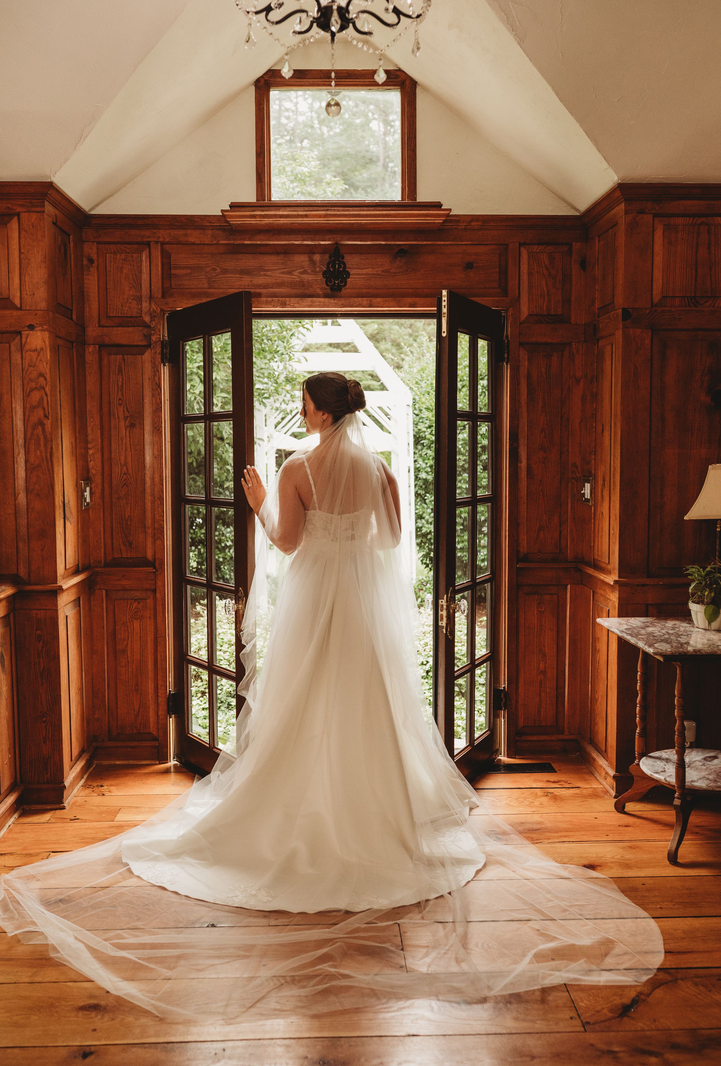 Bride in elegant white wedding dress with long veil standing at open French doors in rustic wood-paneled chapel.