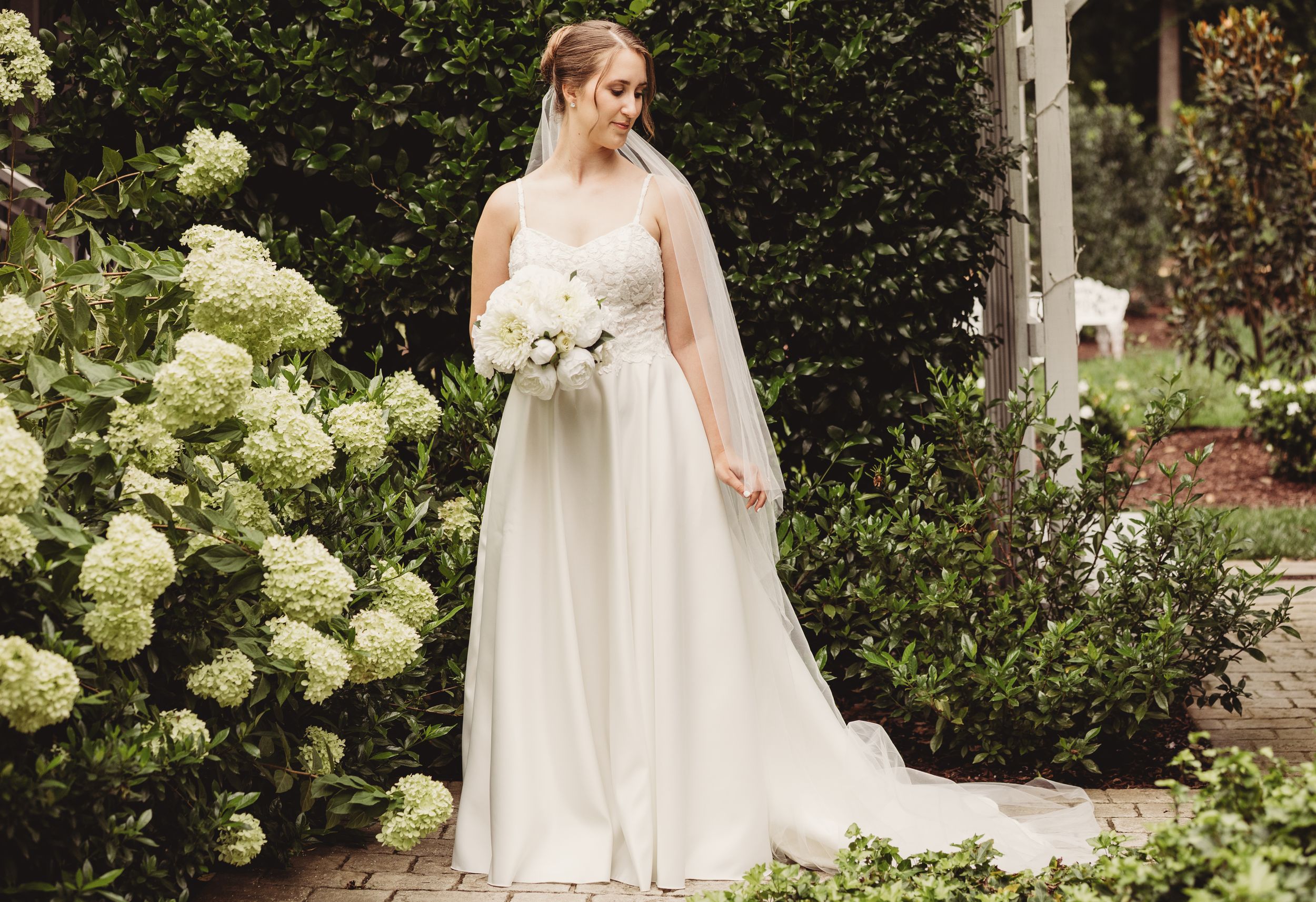 Bride in elegant white wedding gown holding bouquet of white flowers, standing in lush garden with hydrangeas.