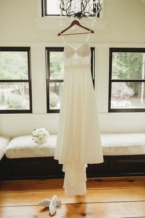 Elegant white lace wedding dress hanging on wooden hanger near window with bridal bouquet and heels below.