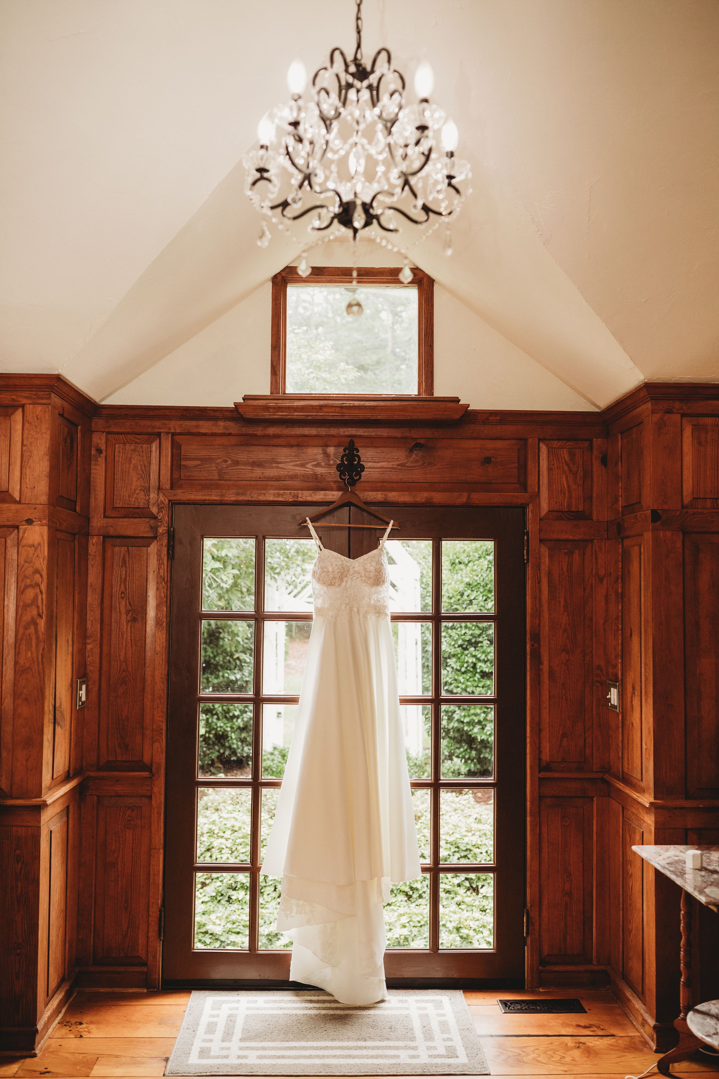White wedding dress hanging on French doors beneath a crystal chandelier in an elegant wood-paneled room.