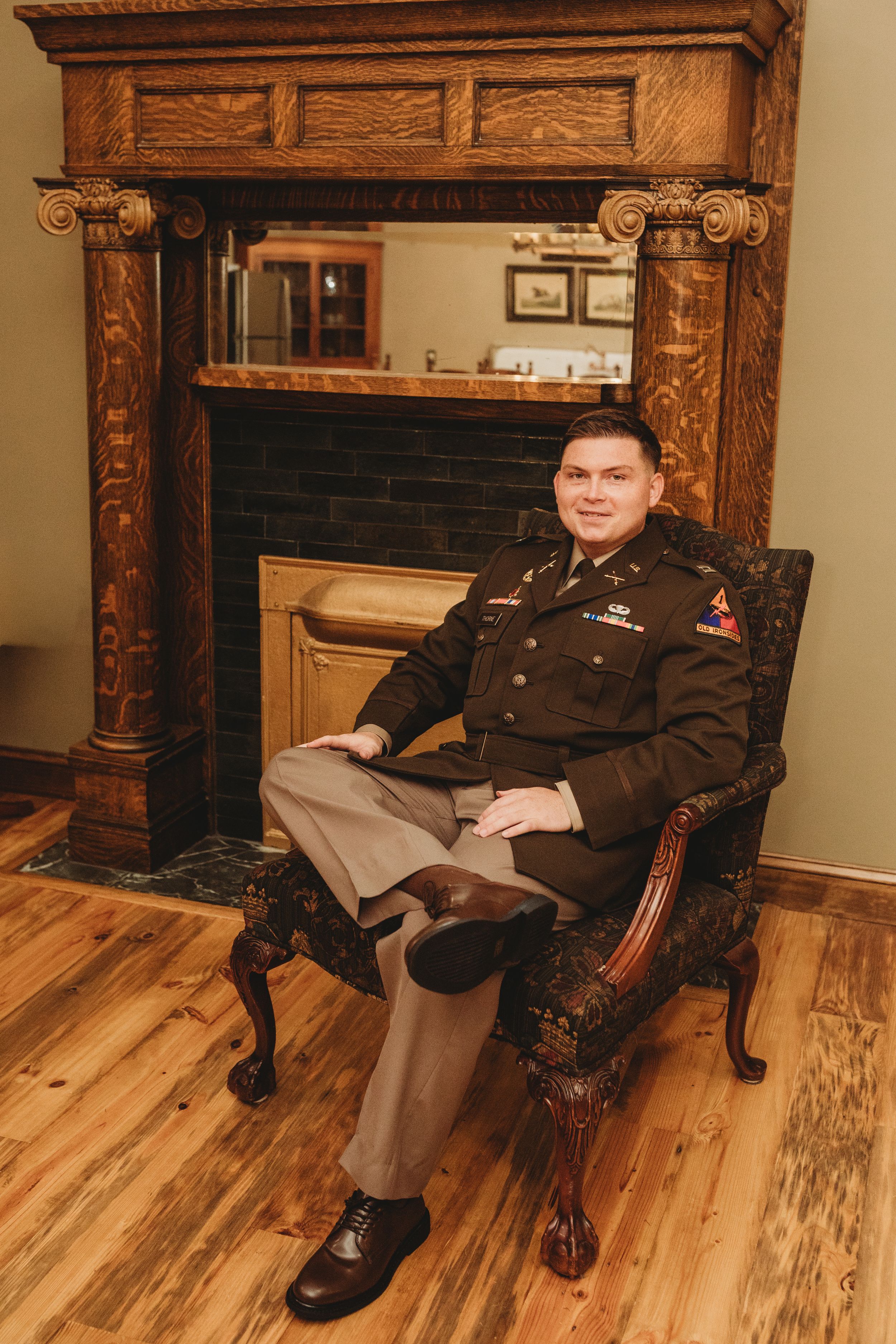 Military officer in dress uniform seated in ornate chair by fireplace.