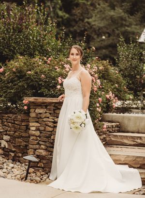 Bride in white lace gown holding bouquet of white flowers, posing in front of stone wall with pink roses.