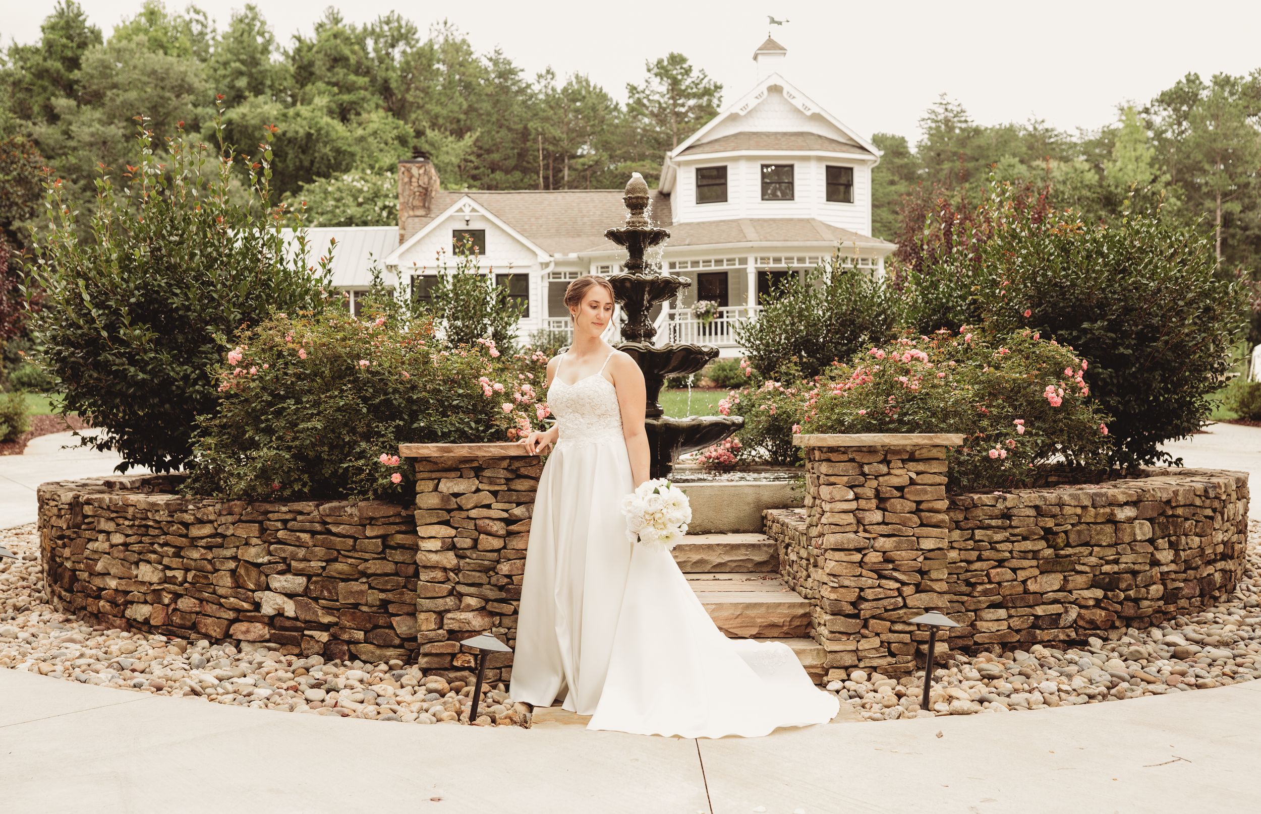 Bride in white gown poses by stone fountain with lush garden and elegant white venue in background.