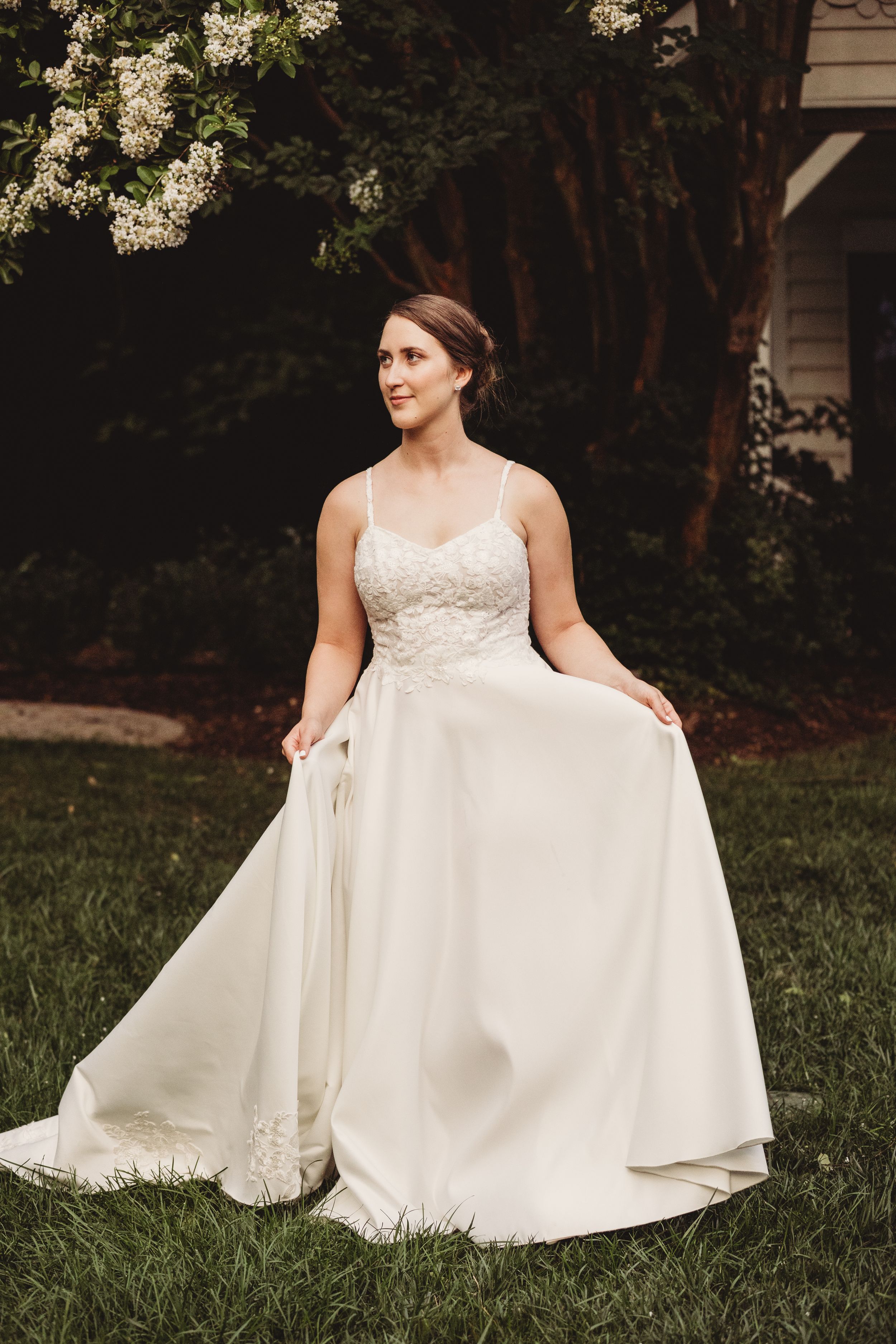 Bride in a white lace spaghetti strap wedding gown holding her skirt, standing on green grass with blooming trees behind her.