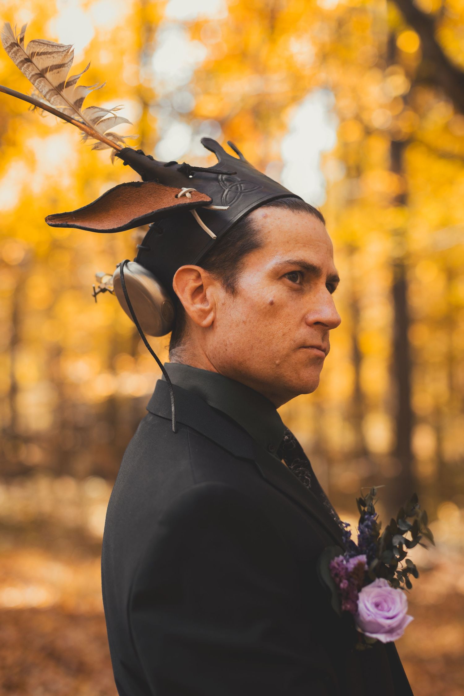 Man in black jacket with decorative headpiece and floral boutonniere poses in autumn forest with golden foliage backdrop.