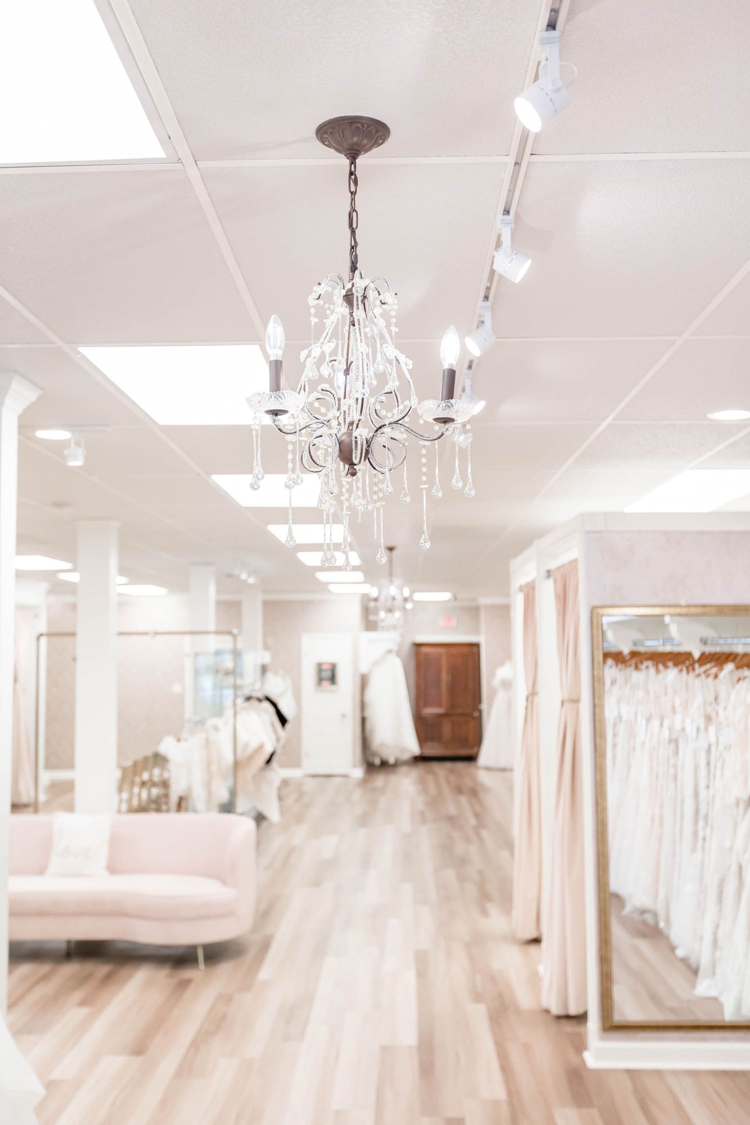 Elegant bridal boutique interior with crystal chandelier, white wedding dresses on racks, and blush pink sofa on hardwood floors.