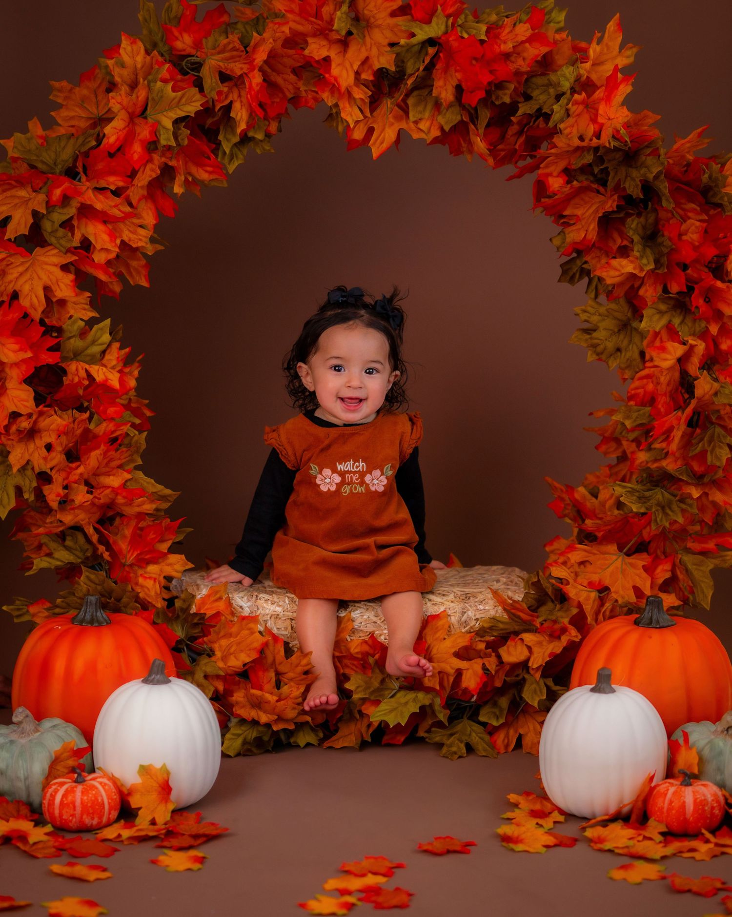 A smiling baby in a rust dress sits among pumpkins and fall leaves under an orange autumn leaf arch.