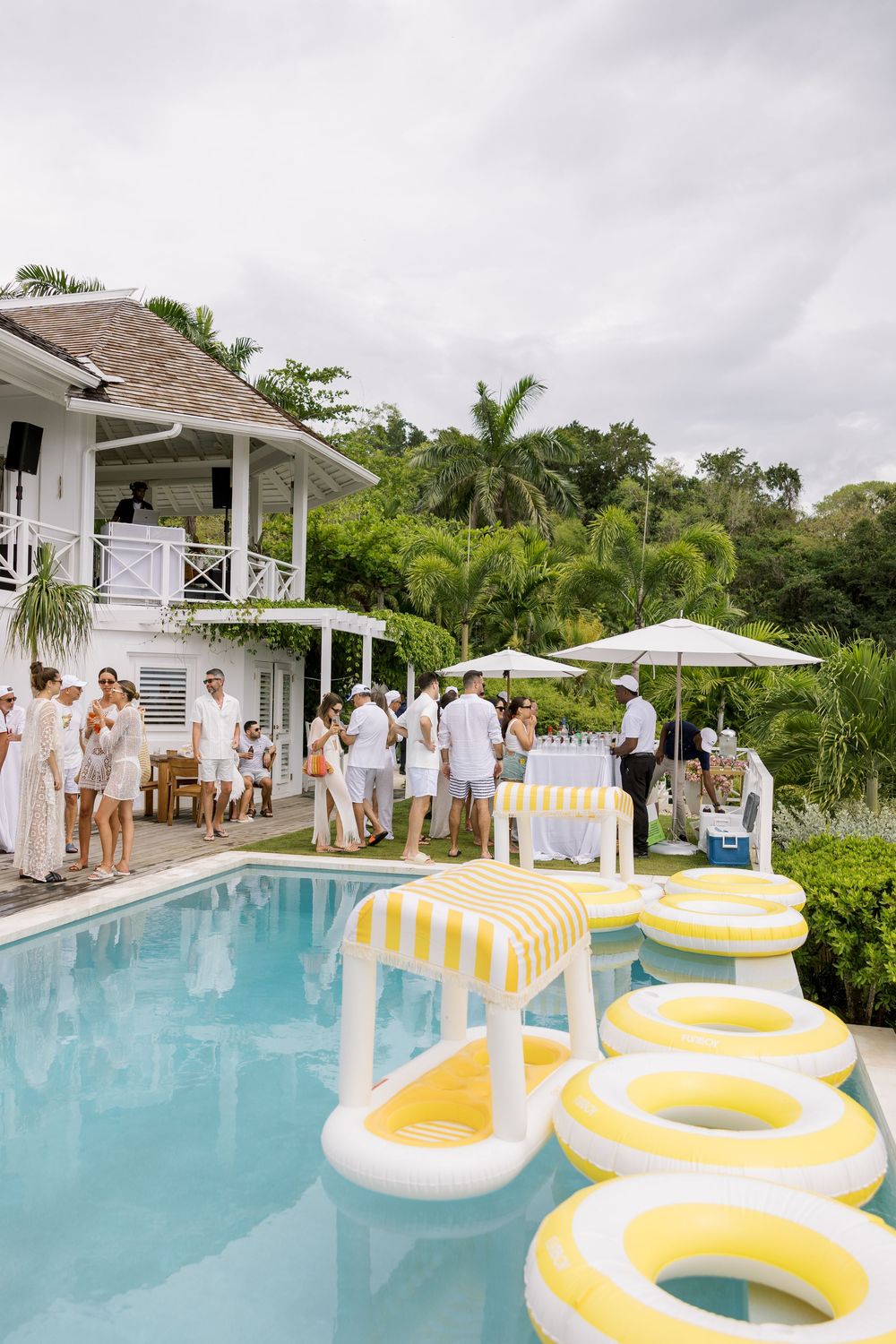 Elegant white party by a pool with yellow float rings, lush tropical greenery, and a white villa in the background.