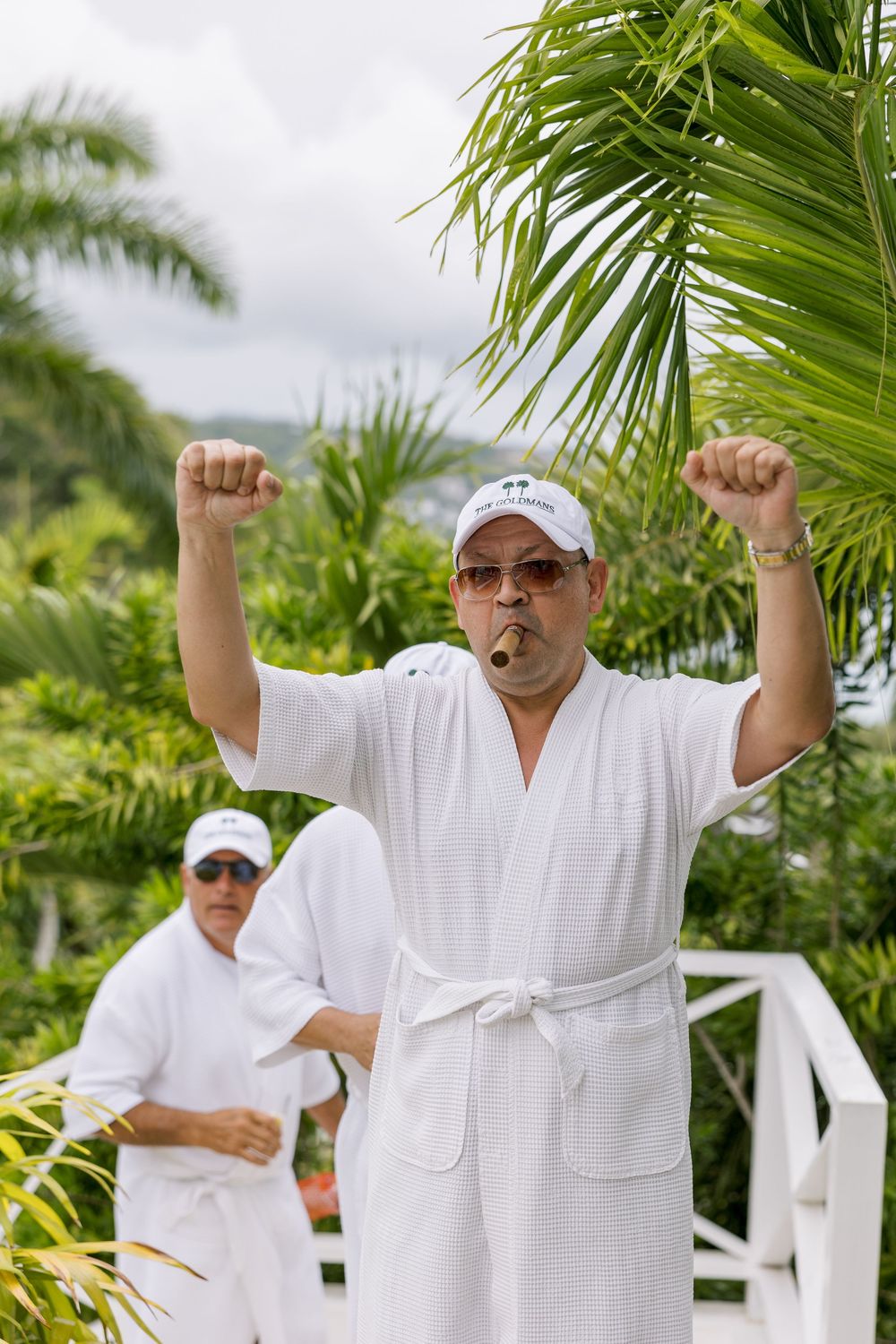 Man in white robe smoking cigar raises fists triumphantly outdoors near tropical palm trees.