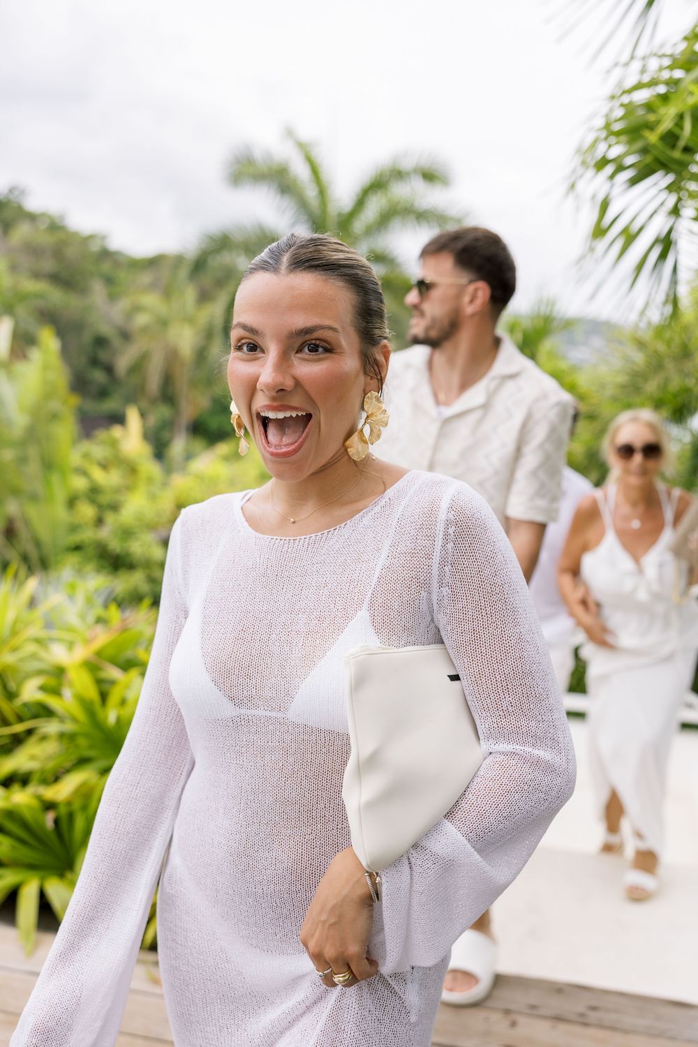 A woman in a white sheer dress with gold earrings smiles excitedly outdoors, with two people in white attire behind her.