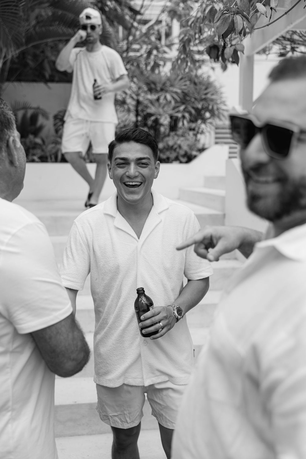 Black and white photo of smiling men in white outfits socializing outdoors with tropical plants in background.
