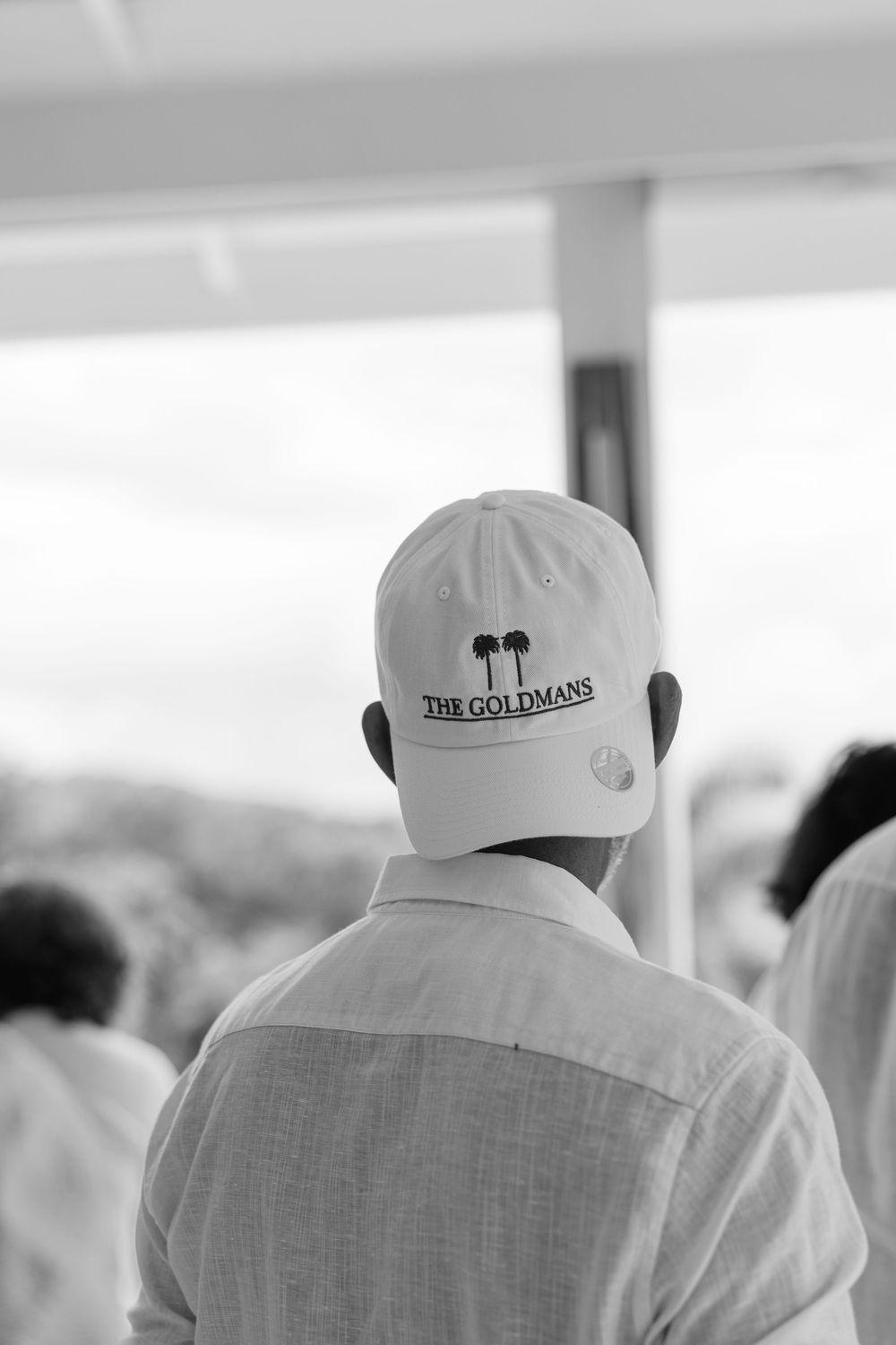 Back view of person wearing white cap reading The Goldmans at an outdoor event in black and white.