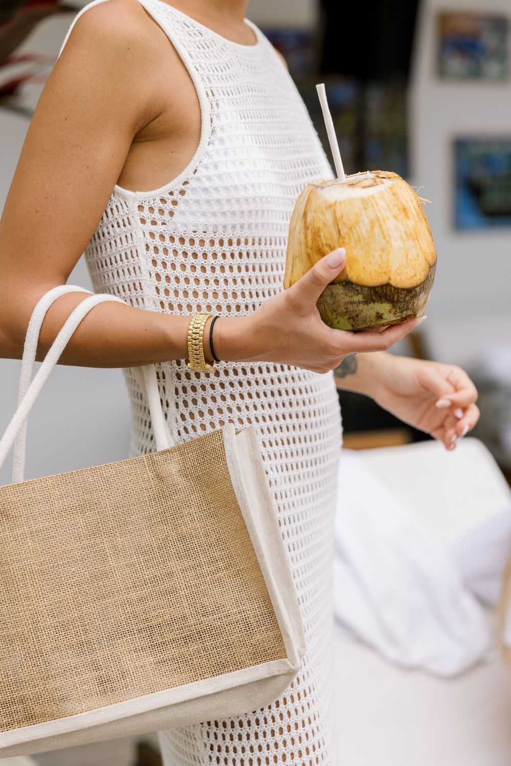 Woman in white crochet dress holding a fresh coconut drink with straw and carrying a woven tote bag.