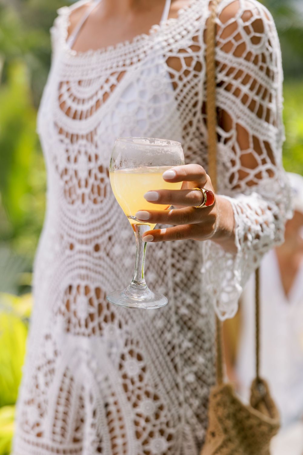 Woman in white crochet dress holding a glass of white wine outdoors in a lush green setting.