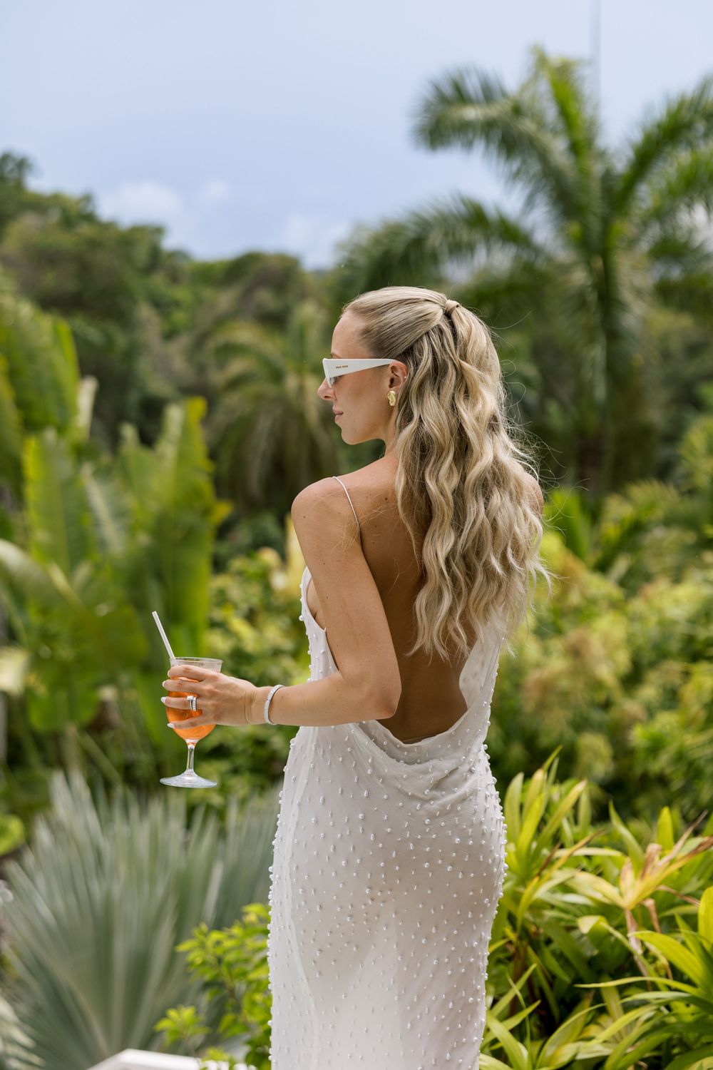 Woman in white backless pearl dress holding cocktail, long blonde wavy hair, tropical garden background.