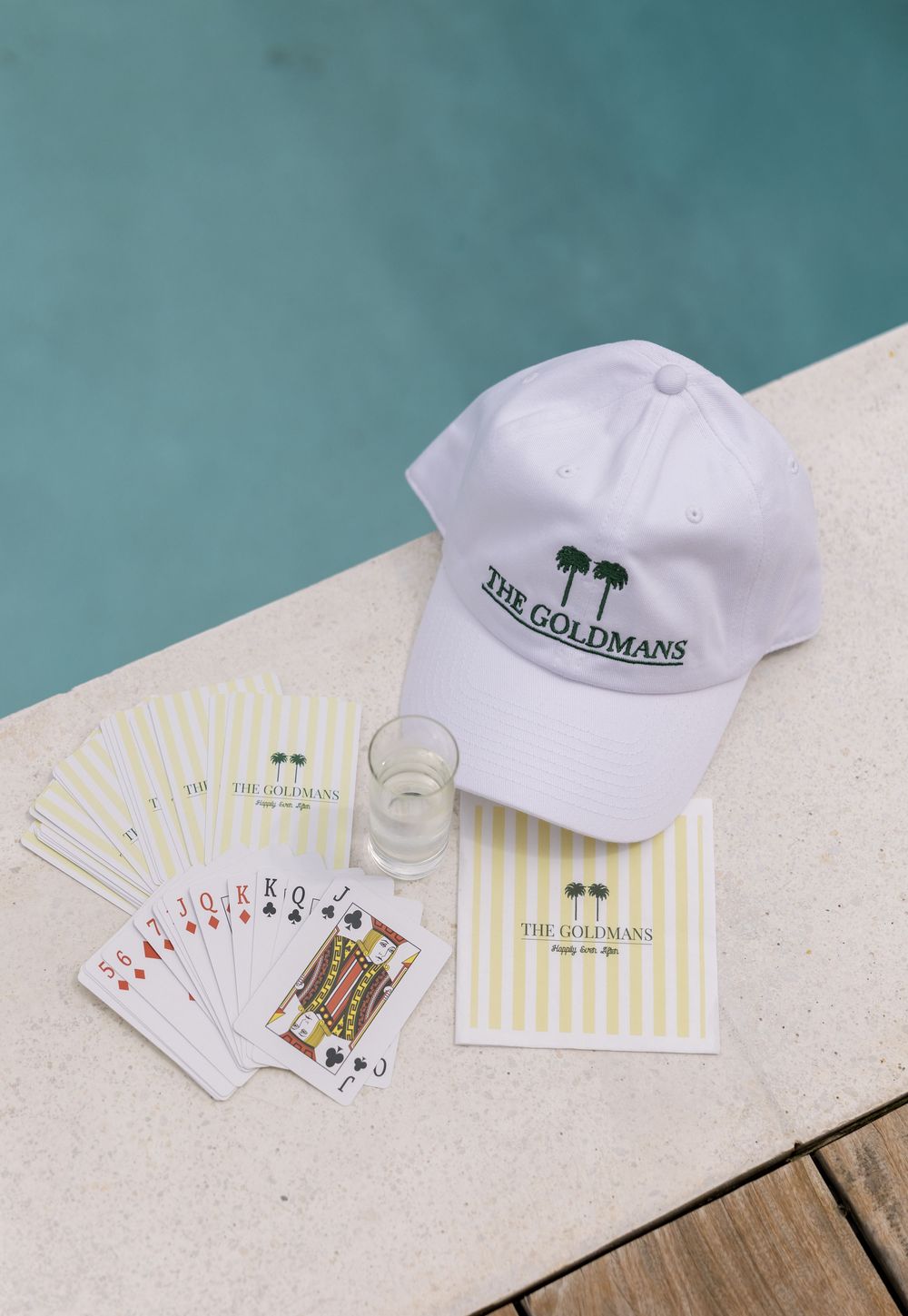 White hat, playing cards, and stationery with The Goldmans palm tree logo displayed poolside.