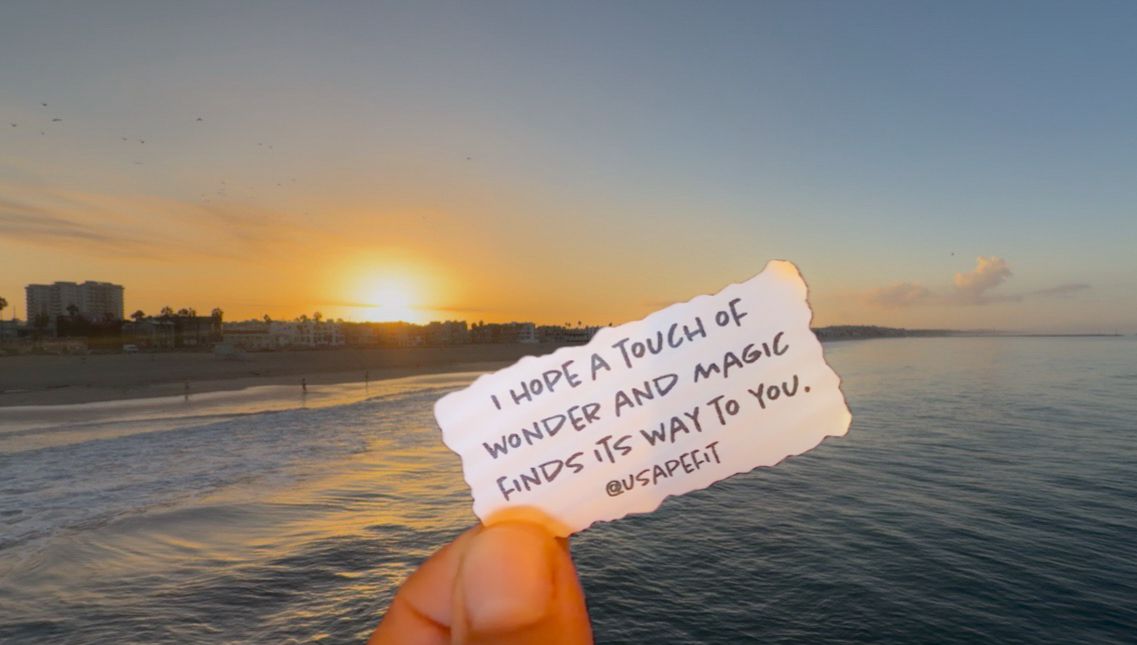 Hand holding a note reading 'I hope a touch of wonder and magic finds its way to you' against a golden sunset beach.