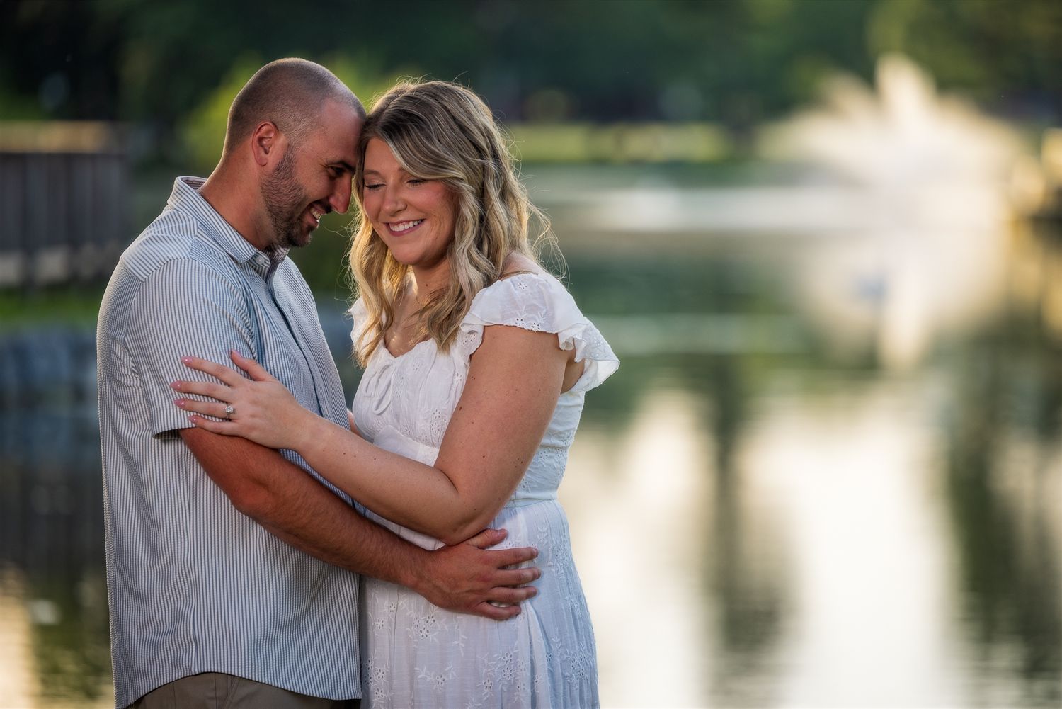 Couple embracing and smiling near a serene pond during golden hour in an outdoor engagement photo session.