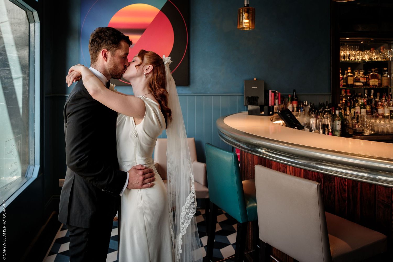 Bride and groom share a romantic kiss near a stylish bar with teal walls and checkered floors.