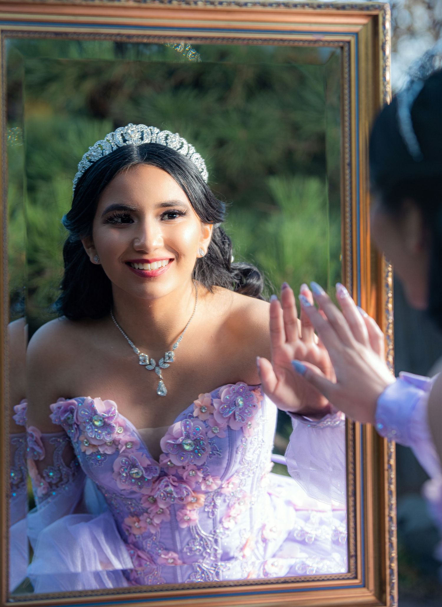 Smiling quinceañera girl in lavender gown and tiara touches a decorative gold mirror outdoors.