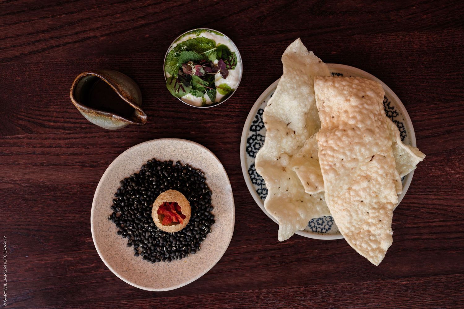 Overhead view of black lentils, flatbread, fresh salad, and sauce on a dark wooden table.