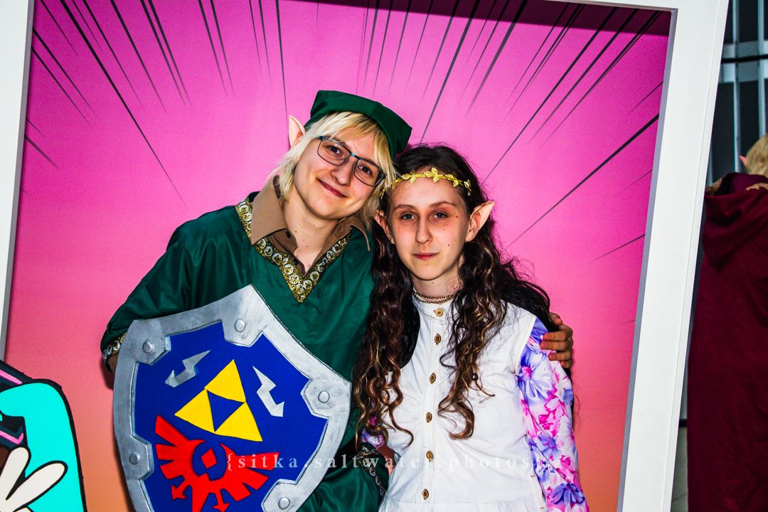 Three cosplayers pose in a decorative anime-themed photo frame at a comic convention.