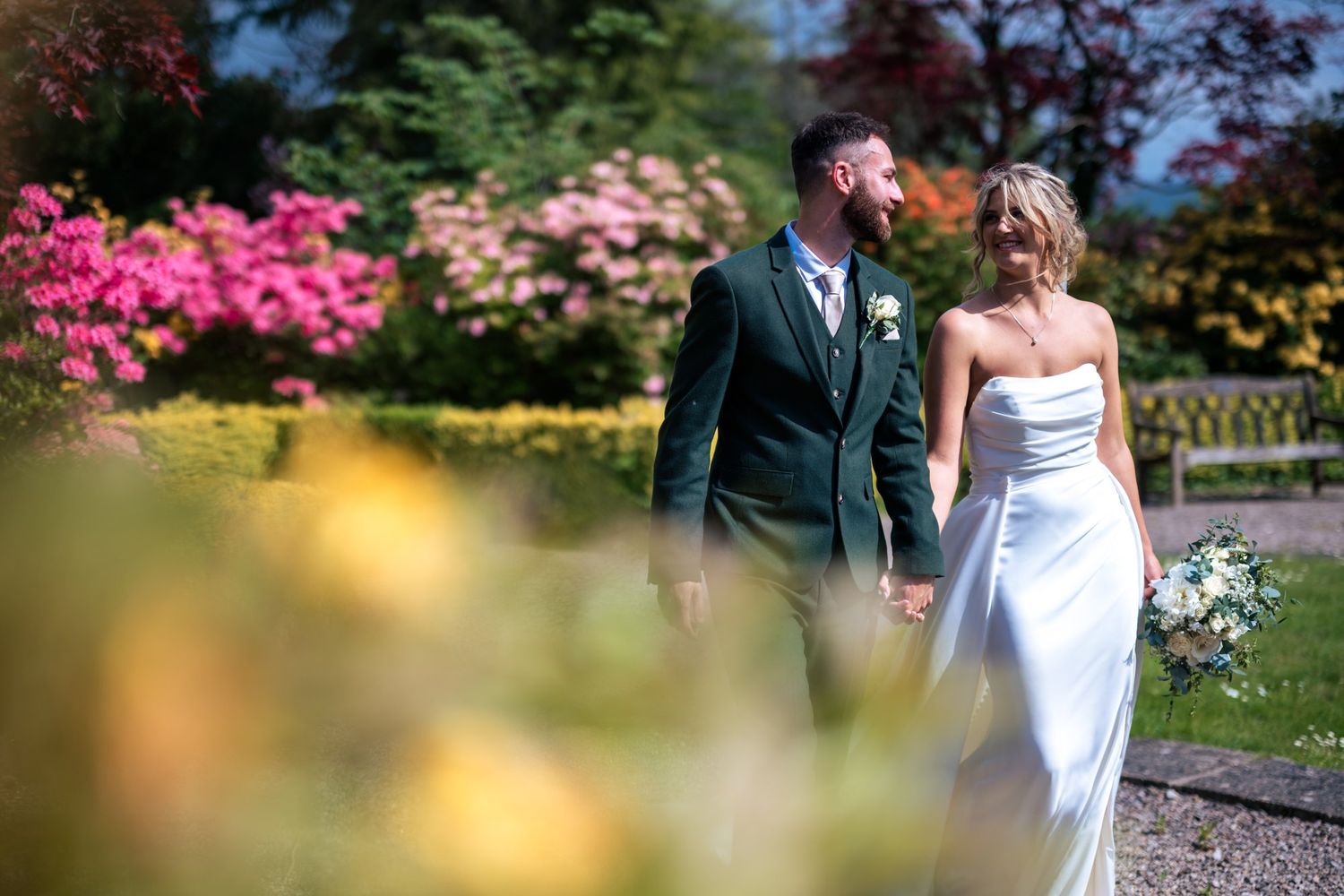 A bride and groom hold hands in a sunlit garden surrounded by vibrant pink and yellow flowers on their wedding day.