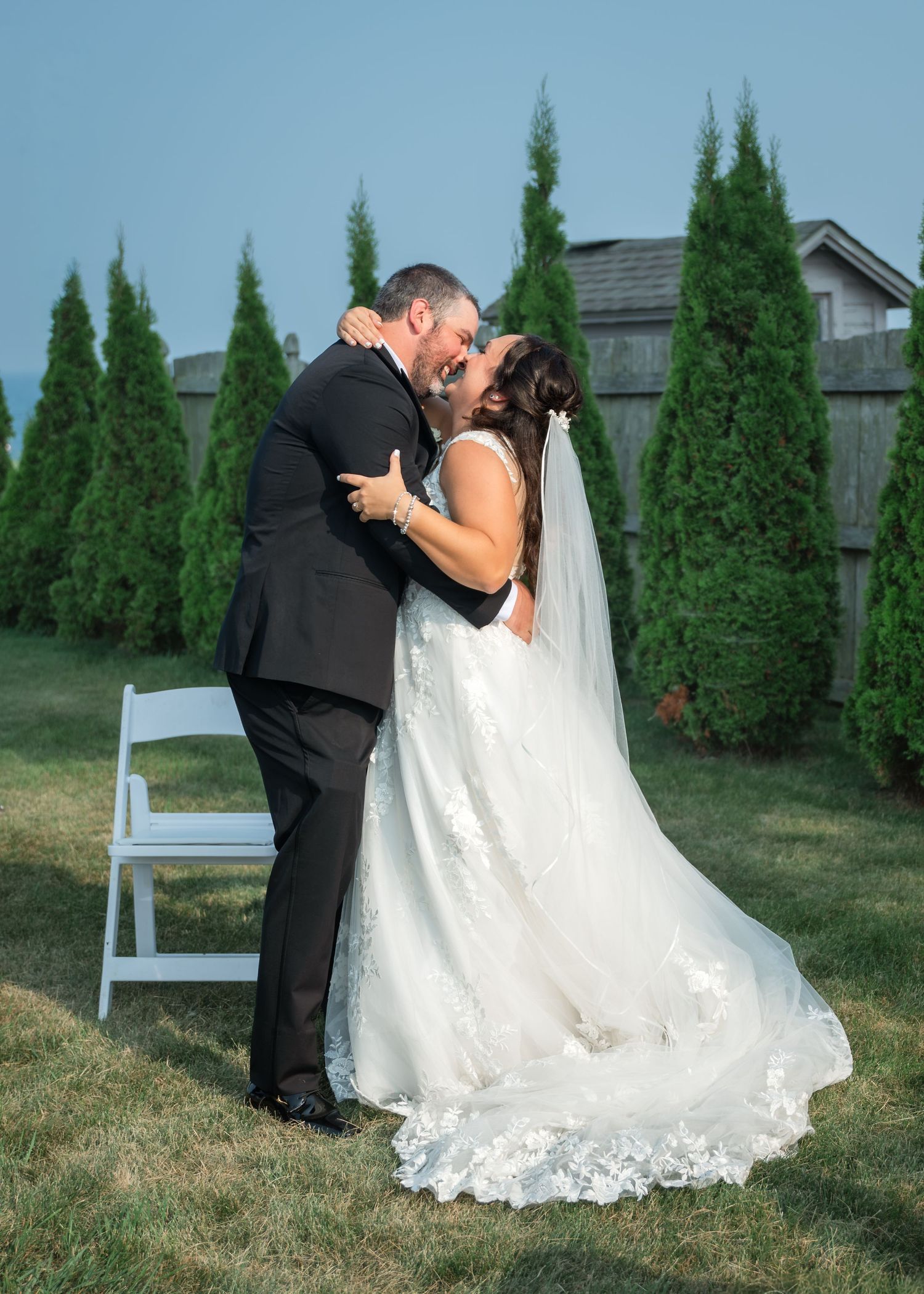 Bride and groom share a romantic kiss outdoors during their wedding ceremony, surrounded by tall evergreen trees.
