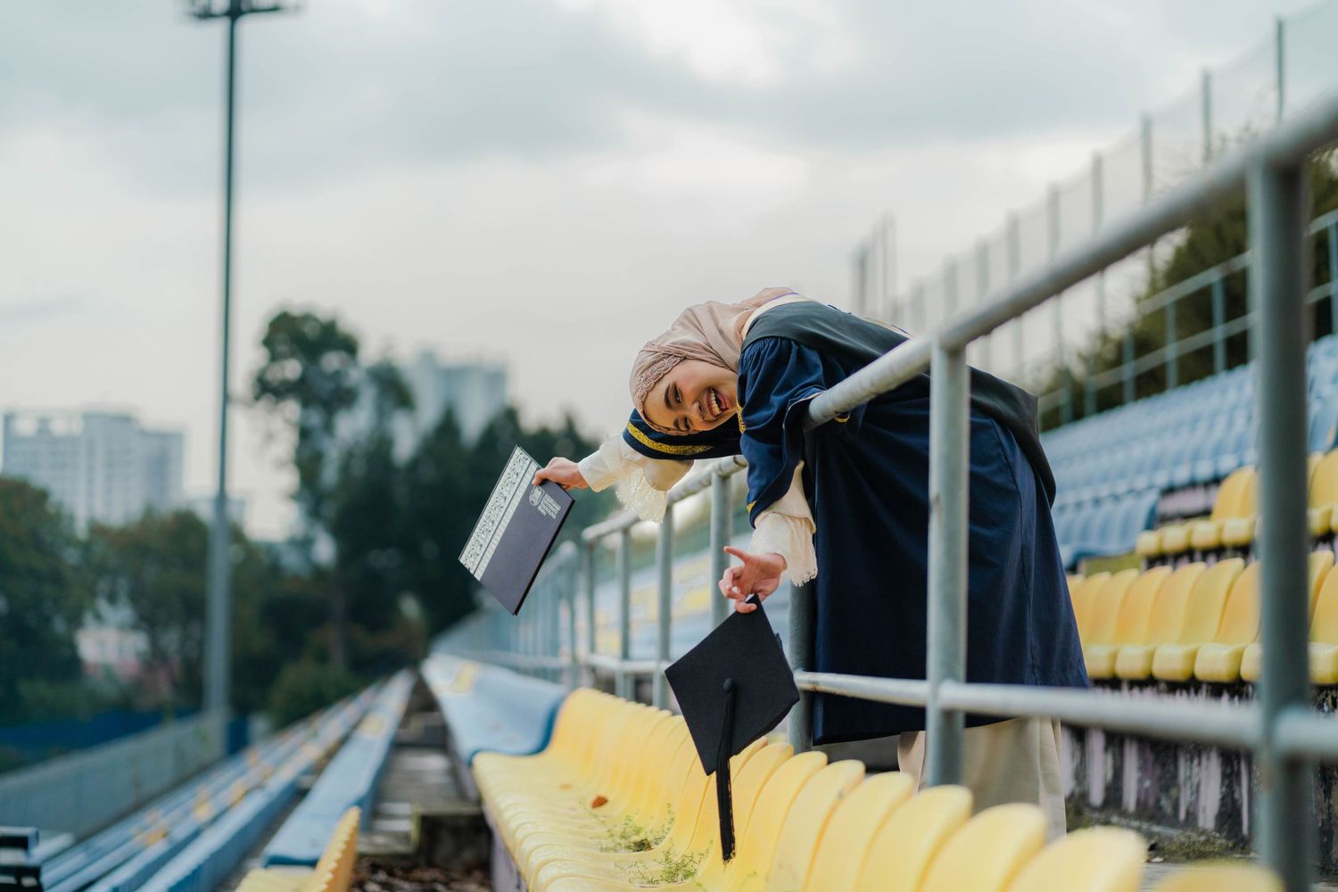 Muslim woman in hijab and graduation gown holding diploma on stadium bleachers, celebrating academic achievement.