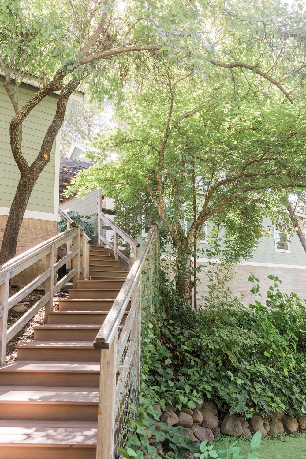 Wooden staircase with railings surrounded by lush green trees leading up to a charming house exterior.
