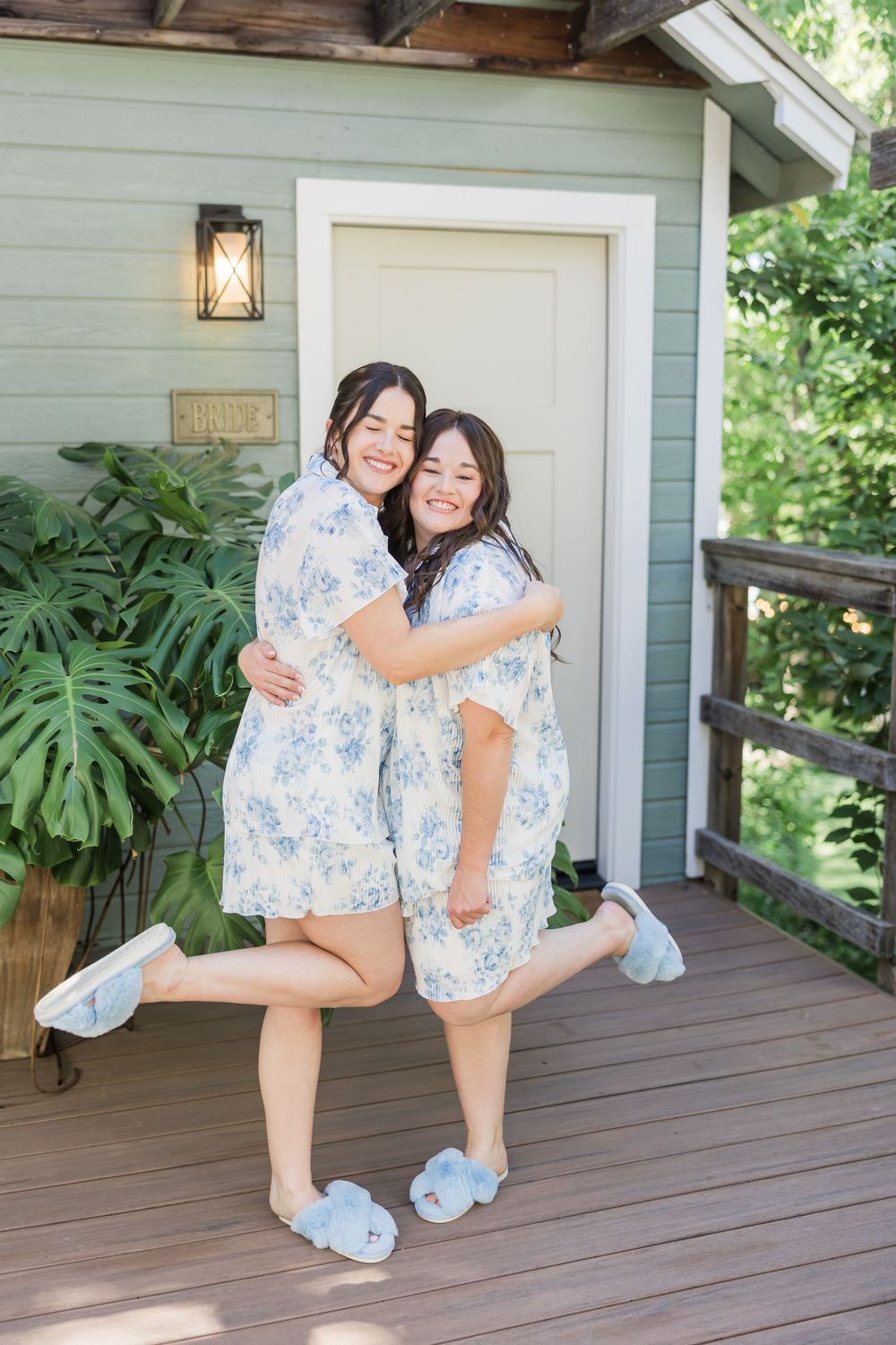 Two women in matching floral blue pajama sets hugging and smiling on a wooden porch of a green cottage.