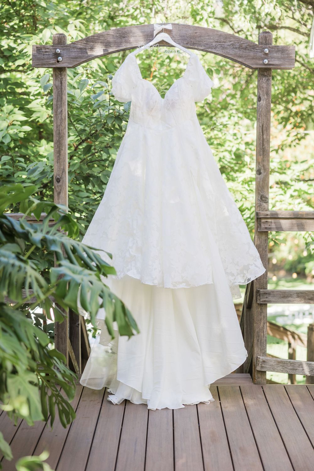 White lace A-line wedding dress with off-shoulder sleeves hanging on wooden arch surrounded by lush green trees.