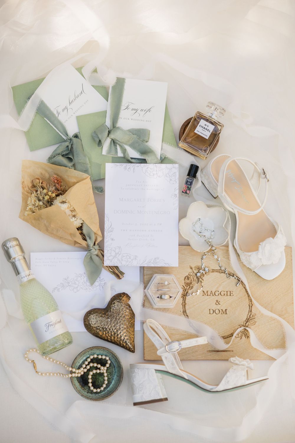 Elegant wedding flat lay with sage green stationery, white heels, perfume, pearls, and personalized wooden sign for Maggie and Dom.