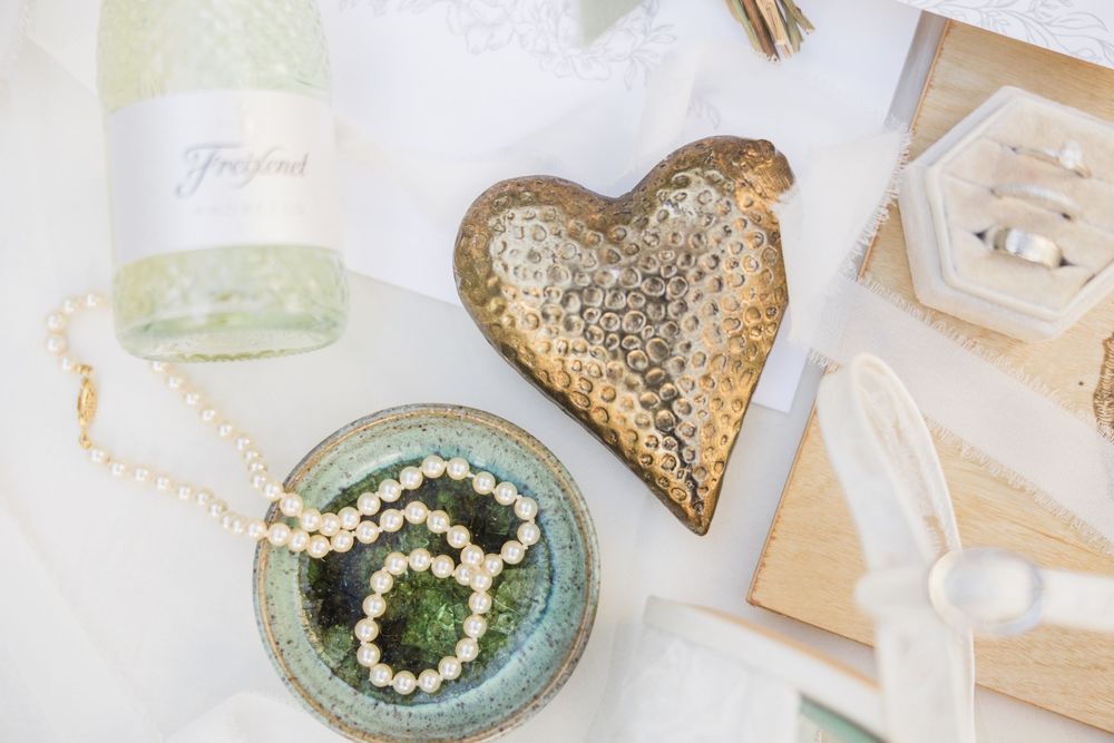 Gold heart dish and pearl bracelet on ceramic plate with wine bottle and wooden accents in elegant flat lay.