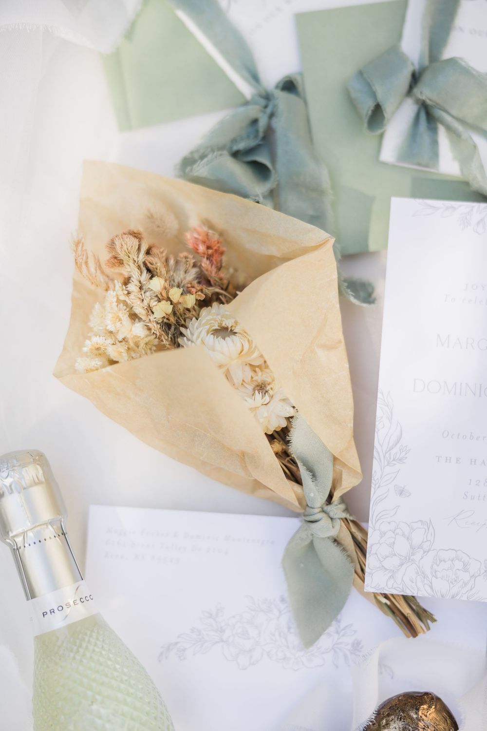 Dried flower bouquet wrapped in kraft paper with sage green ribbon beside wedding stationery and Prosecco.