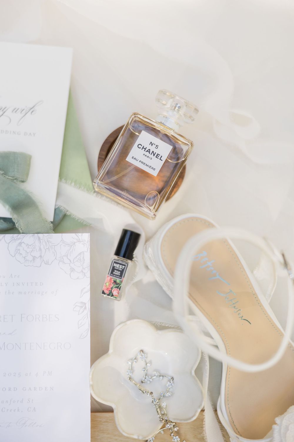 Chanel No. 5 perfume, white bridal heels with floral detail, wedding stationery, and a small perfume roller on white fabric.