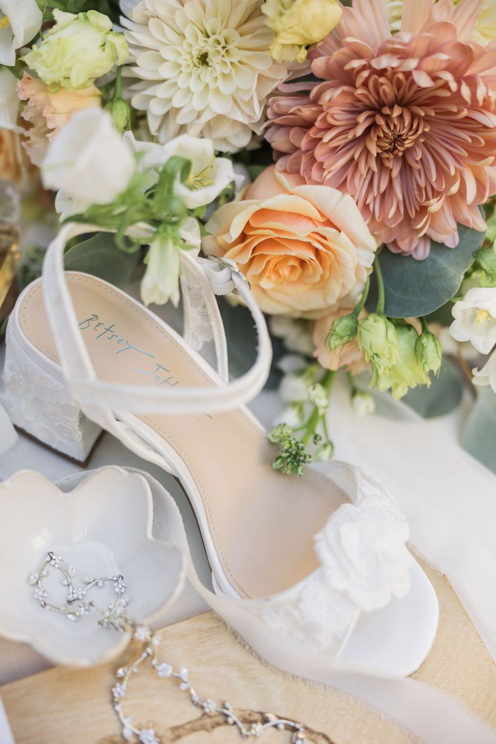 Elegant white bridal heels with floral detail beside a lush bouquet of dahlias, roses, and crystal jewelry.