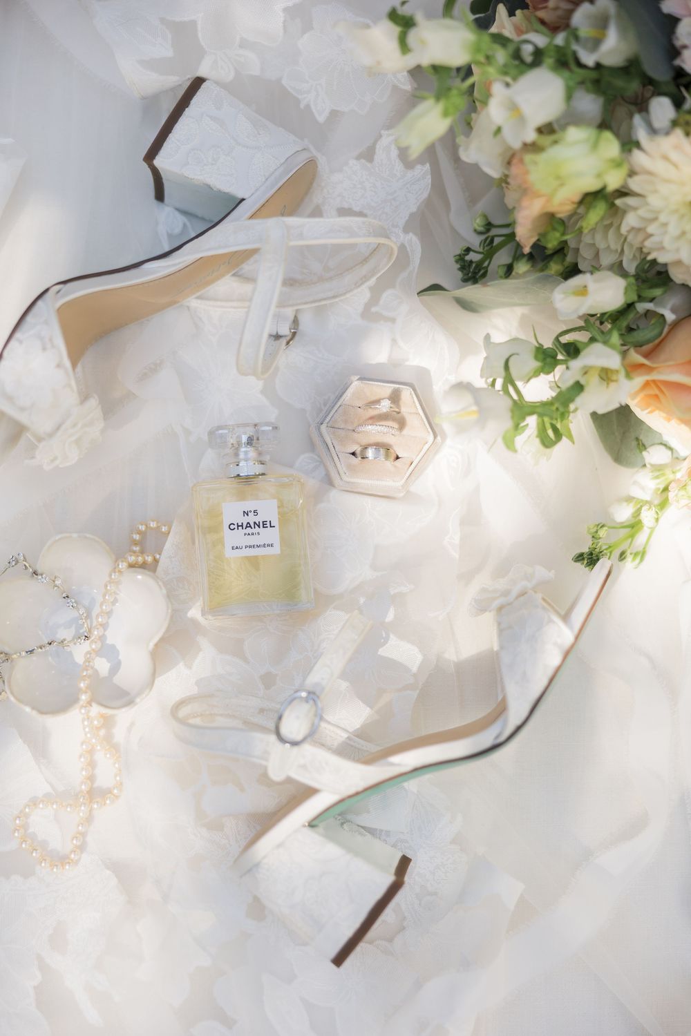 Bridal flat lay with Chanel No.5 perfume, white heels, pearl necklace, wedding rings in velvet box, and floral bouquet.
