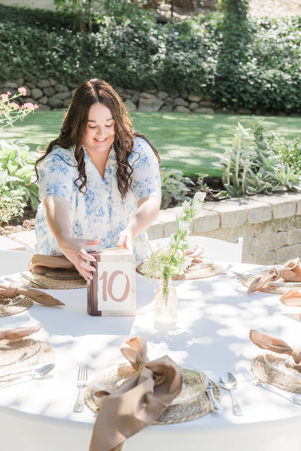 Woman arranging table number 10 at an elegant outdoor wedding reception with woven chargers and floral centerpieces.