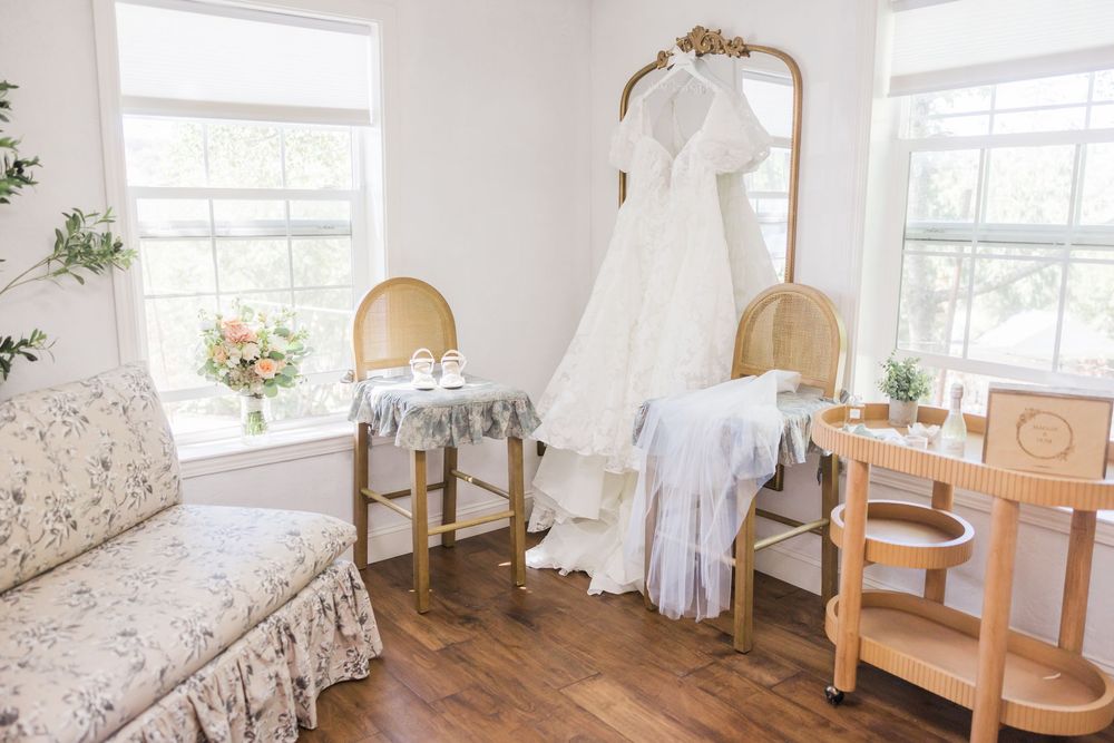 Elegant bridal suite with wedding dress hanging on ornate gold mirror, floral bouquet, and vintage chairs on hardwood floors.